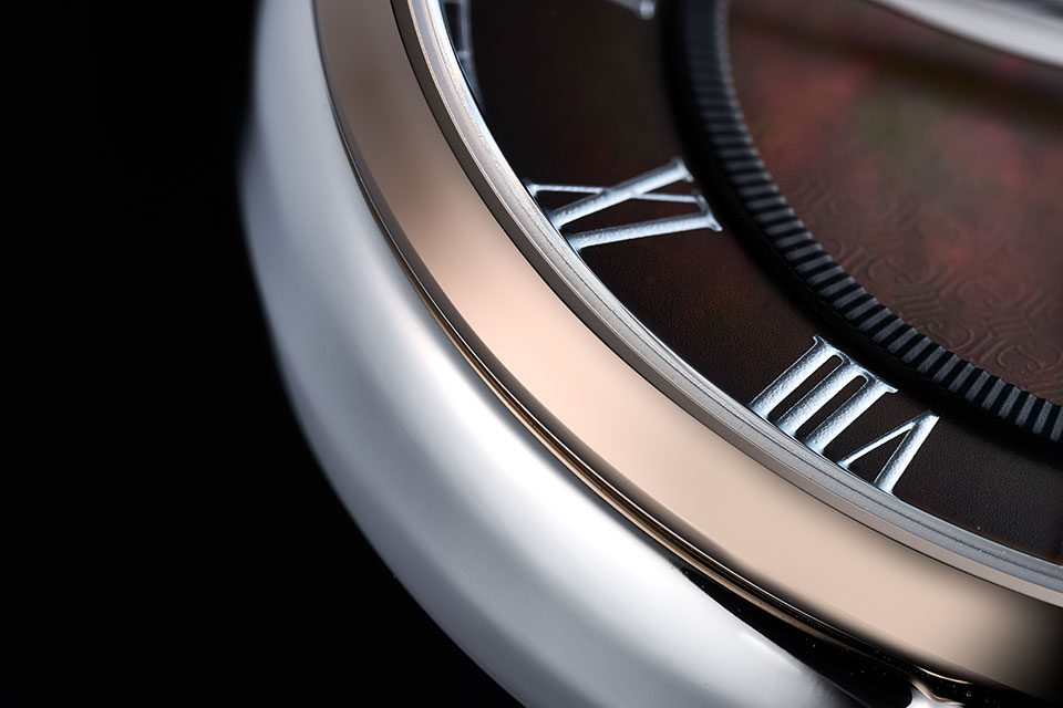ブラウンのグラデーションダイアルとの連動性を持たせるべく、ベゼルにはブロンズ色のめっきが施される。繊細かつ立体的なローマンインデックスにはタコ印刷の技術が用いられ、時計全体をエレガントな雰囲気に仕上げている。
