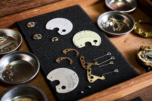 工房の机に並べられた「6号」のパーツ。レトログラードの分・時スケールの字入れ段階別のダイアルや、2種類のレトログラード針、カム等が並ぶ。